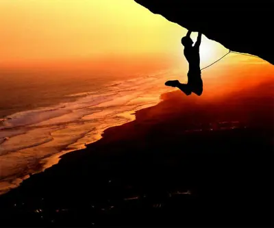 The silhouette of a free-climbing man attempting to pull himself over the edge of a cliff as he dangles over thin air. There is a bright-orange sunset behind him.