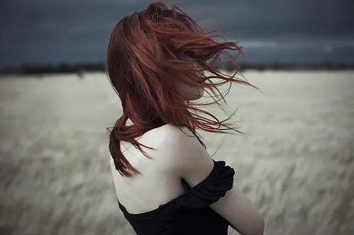 A red-haired woman standing in a wheat field, facing away from the viewer. Her hair blows foward, obscuring her face. A strap of her dress slips off her pale shoulder.
