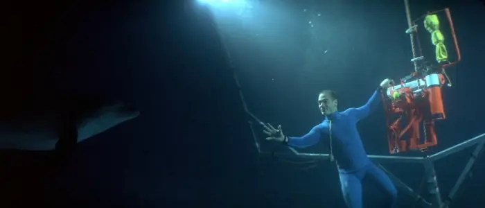 Jack in his blue free-diving suit, more than 100 feet underwater where it is pitch-black. He holds to a riging platform that is equipped with flood lights. Just out of reach of the light is a dolphin, looking at him.